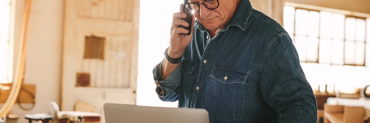 A man making a phone call while looking at his laptop in a sunny workshop.