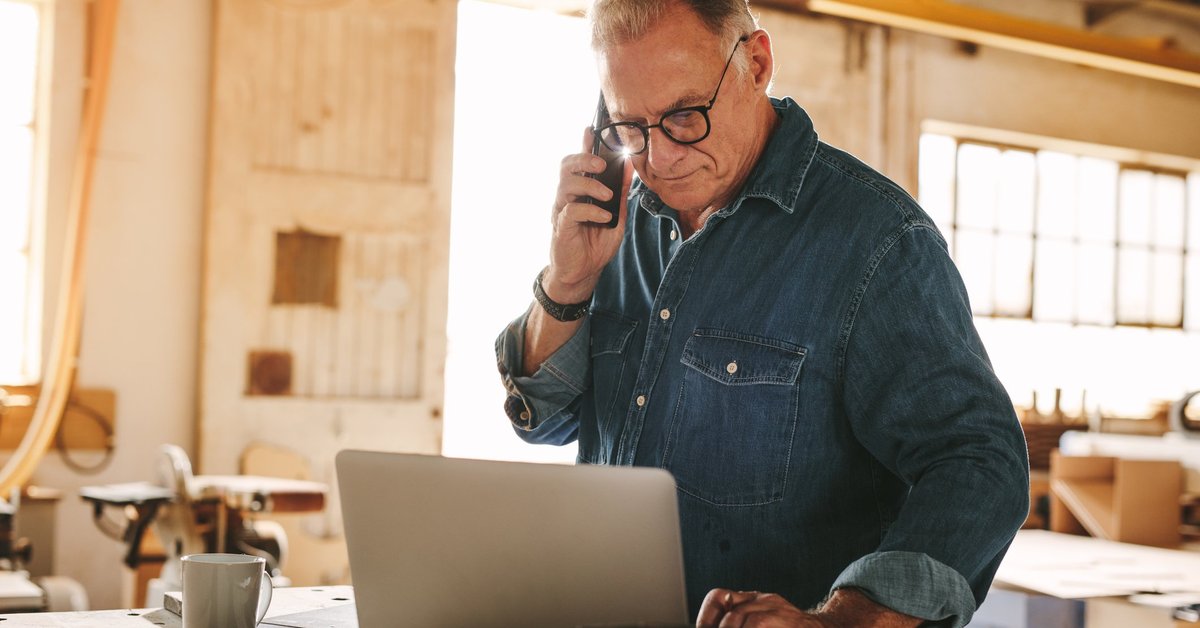A man making a phone call while looking at his laptop in a sunny workshop.