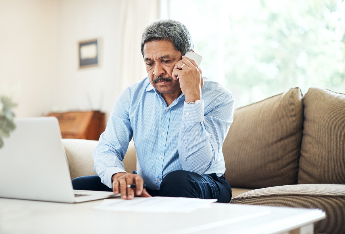 An older man looking annoyed on a phone call while sitting on his couch with his laptop on the table in front of him.