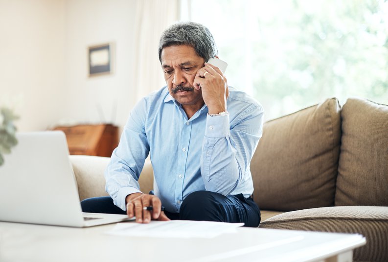 An older man looking annoyed on a phone call while sitting on his couch with his laptop on the table in front of him.