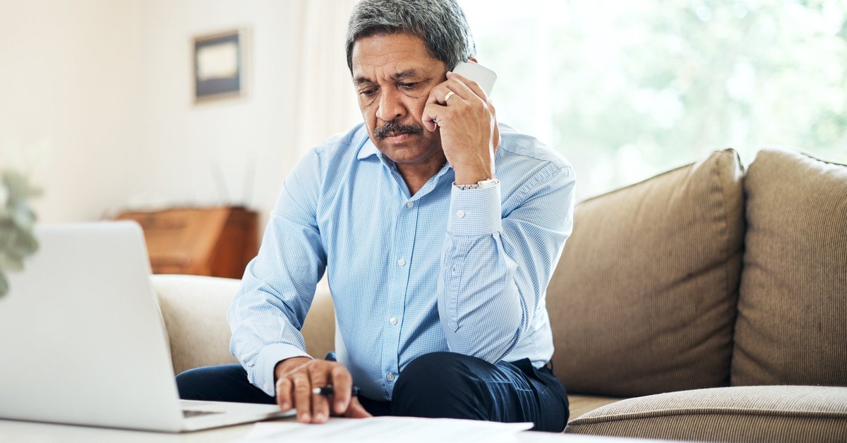 A man on a phone call while sitting on his couch with his laptop in front of him.