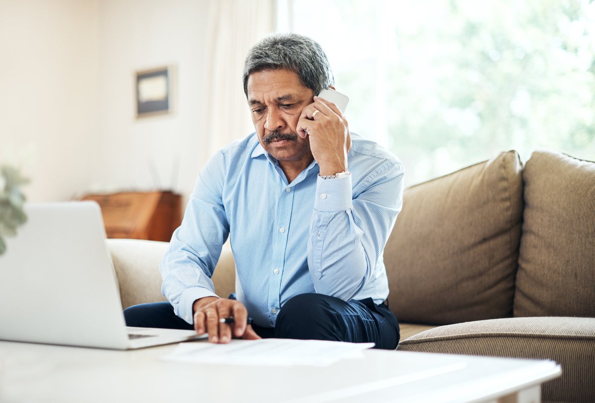 A man talks on the phone while sitting on his couch with his laptop in front of him.