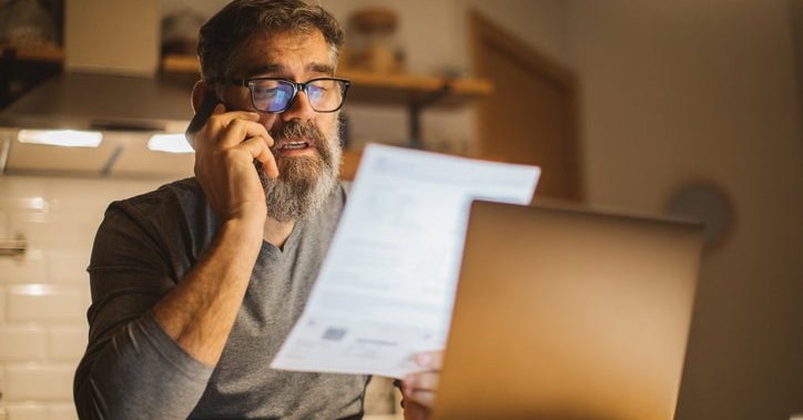 A man sitting at a desk in front of a laptop and making a phone call while holding up and reading a piece of paper.