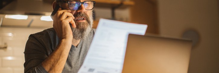 A man sitting at a desk in front of a laptop and making a phone call while holding up and reading a piece of paper.