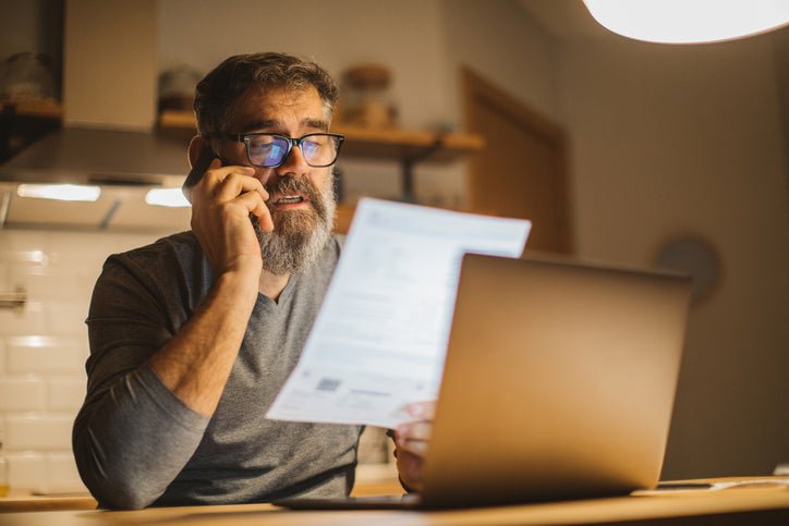 A man sitting at a desk in front of a laptop and making a phone call while holding up and reading a piece of paper.