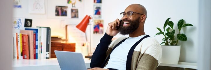 A smiling man on a phone call sitting with his laptop on his lap while working from his home office.