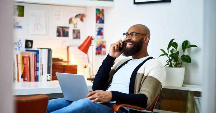 A smiling man on a phone call sitting with his laptop on his lap while working from his home office.