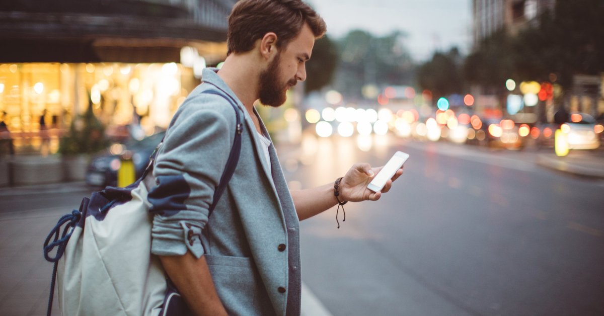 A man standing on a street corner at dusk and looking at his phone.