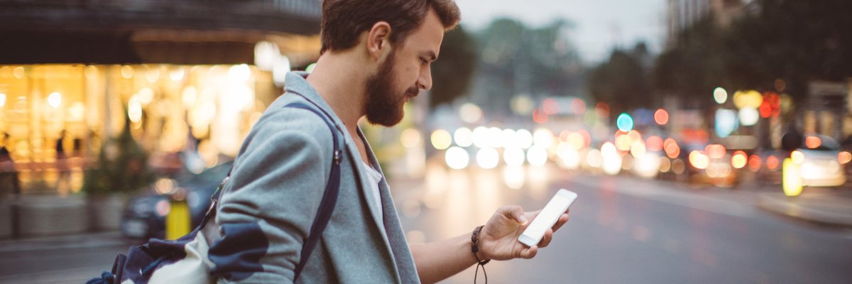 A man standing on a street corner at dusk and looking at his phone.