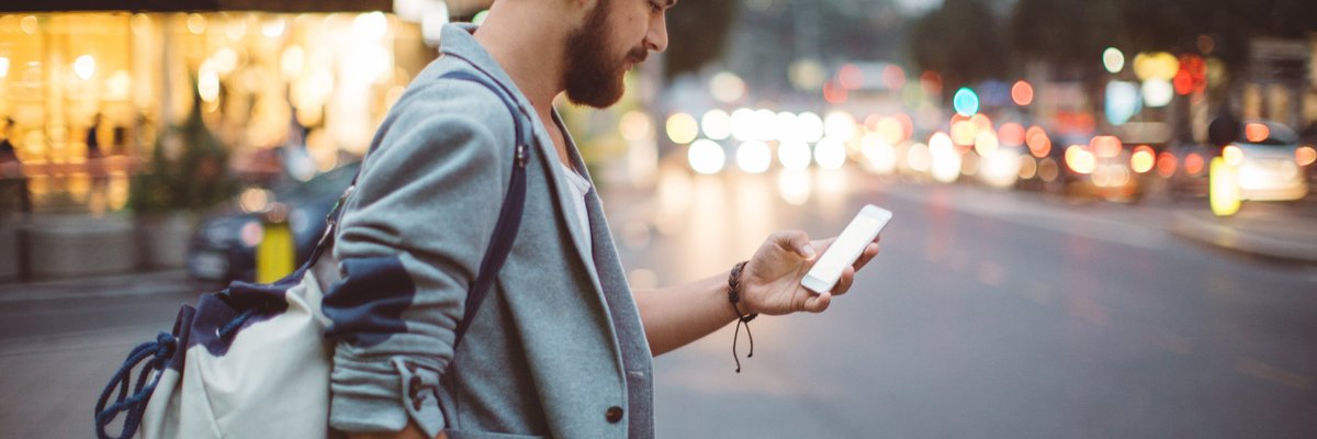 A man standing on a street corner at dusk and looking at his phone.