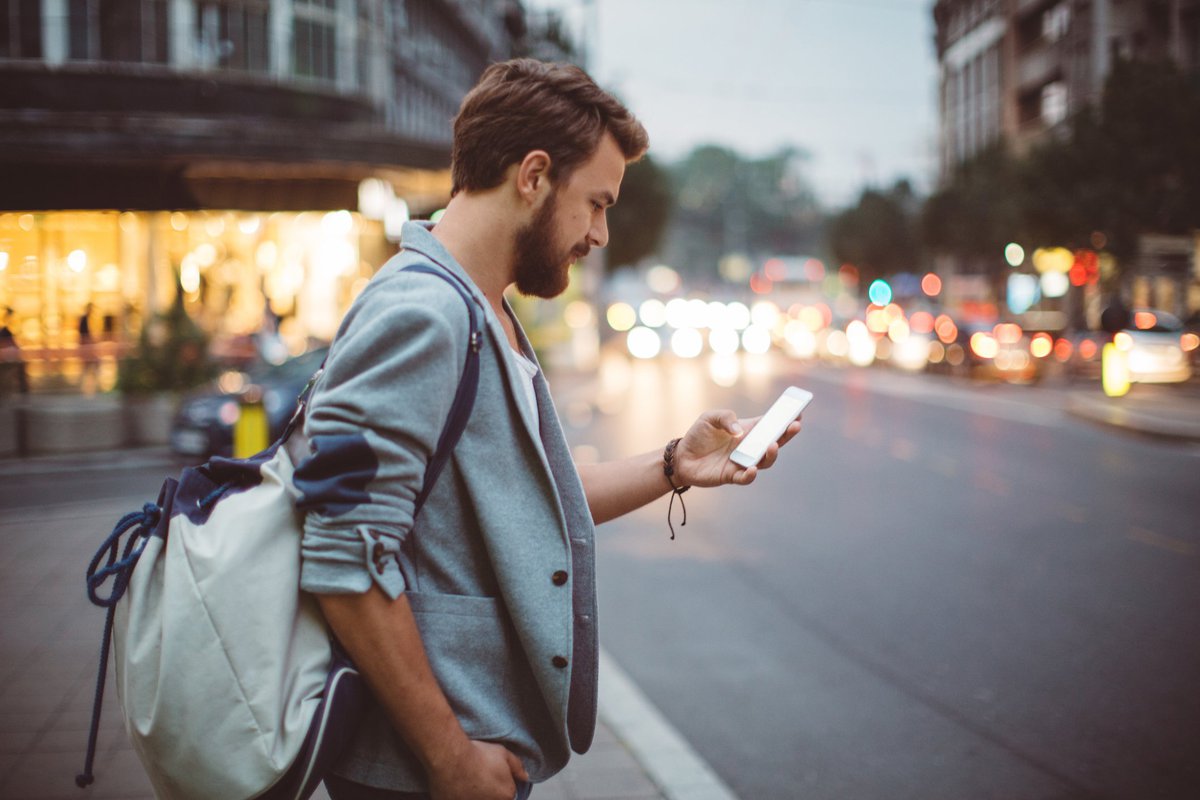 A man standing on a street corner at dusk and looking at his phone.