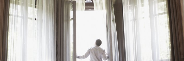 A man opening the floaty curtains and floor-to-ceiling glass balcony doors in a nice hotel room.