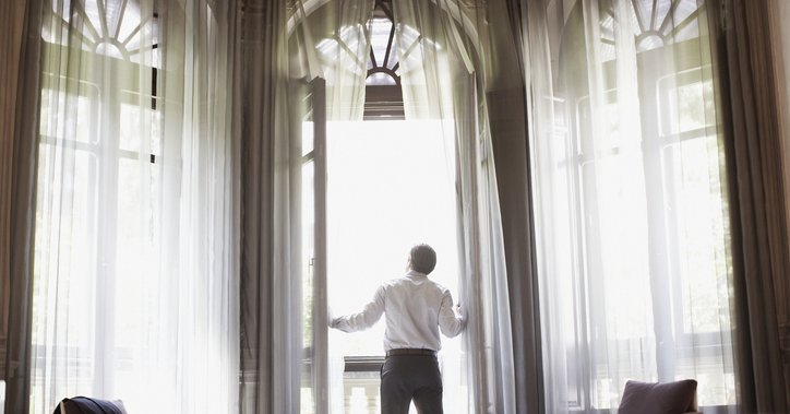 A man opening the floaty curtains and floor-to-ceiling glass balcony doors in a nice hotel room.