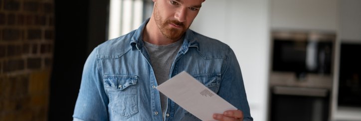 A man standing in his living room and holding a pile of mail in one hand while looking at a single envelope in the other hand.