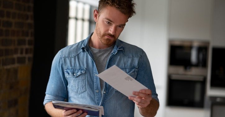 A man standing in his living room and holding a pile of mail in one hand while looking at a single envelope in the other hand.