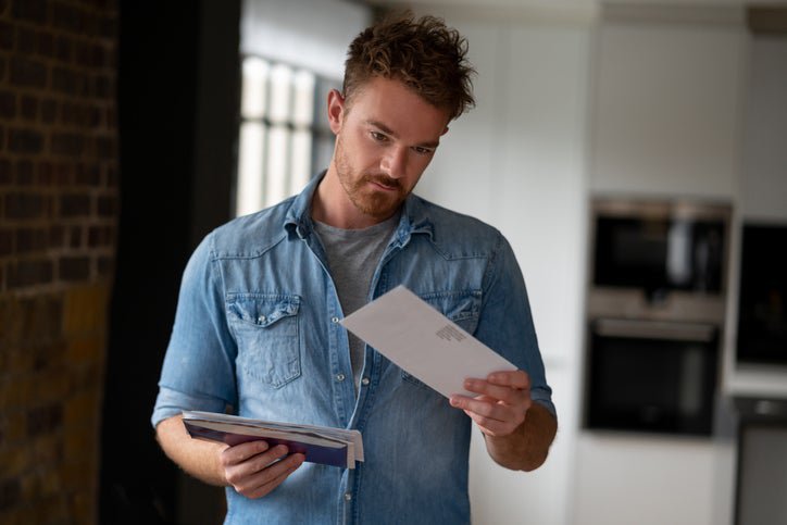 A man standing in his living room and holding a pile of mail in one hand while looking at a single envelope in the other hand.