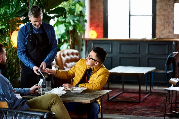 A man sitting at a table in a casual restaurant and tapping his credit card on the payment reader being held by the waiter.