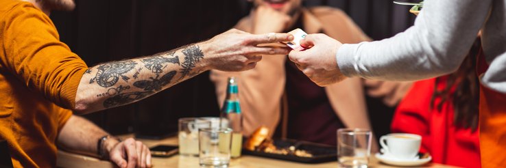 A man handing his credit card to a waiter at a restaurant while sitting at a table with his friend.