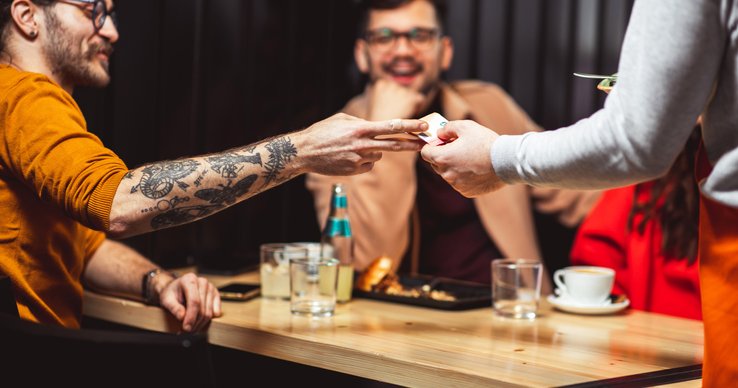 A man handing his credit card to a waiter at a restaurant while sitting at a table with his friend.