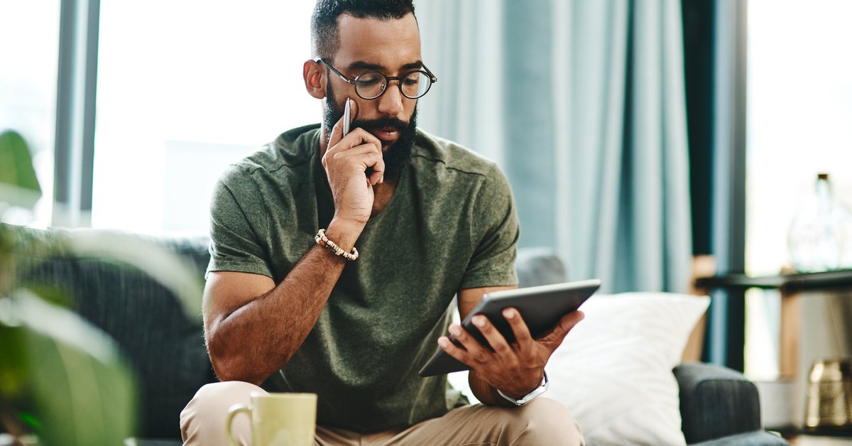 A man reviewing his finances on a tablet with bills on the coffee table in front of him.