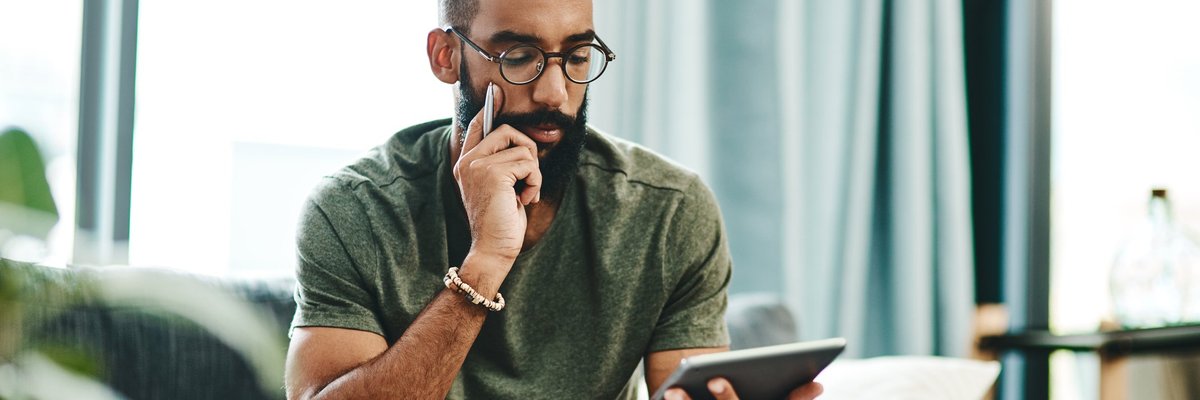 A man reviewing his finances on a tablet with bills on the coffee table in front of him.