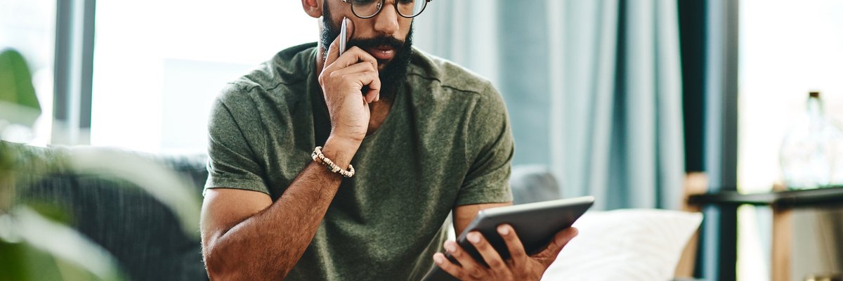 A man reviewing his finances on a tablet with bills on the coffee table in front of him.