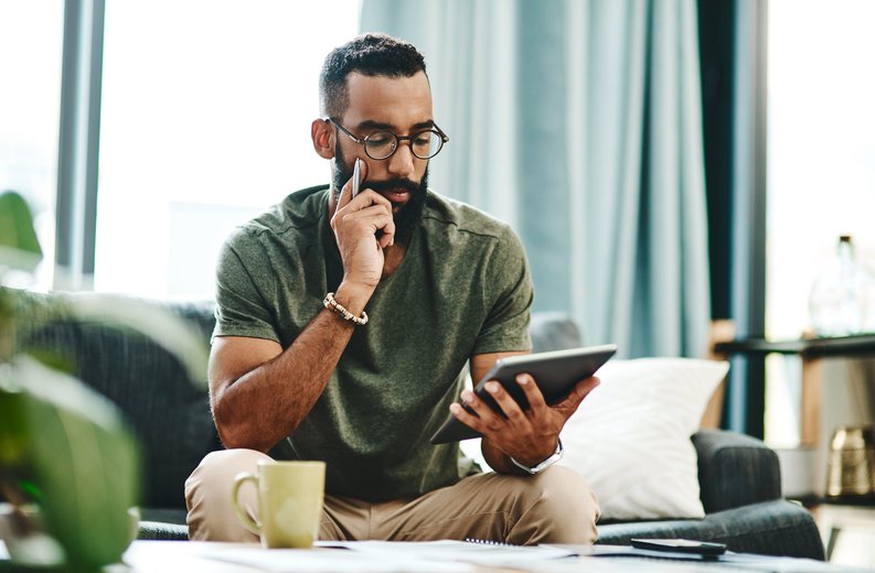 A man reviewing his finances on a tablet with bills on the coffee table in front of him.