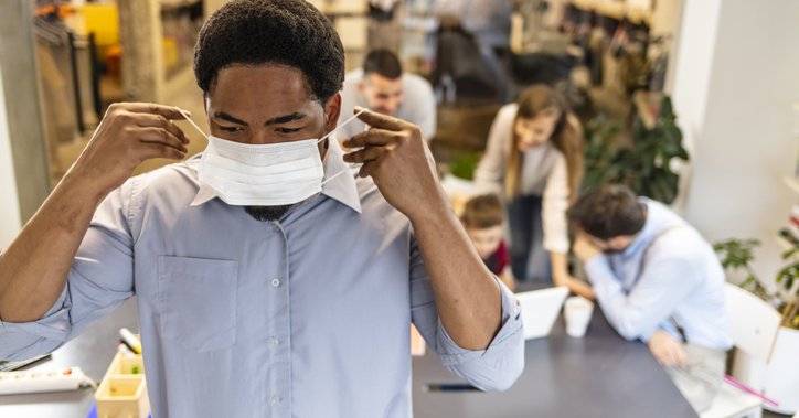 A man putting on a medical mask with his coworkers talking at a table behind him.