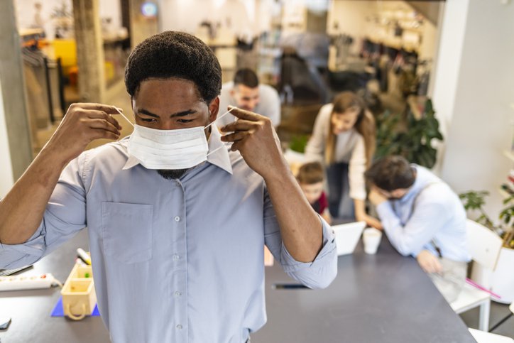 A man putting on a medical mask with his coworkers talking at a table behind him.