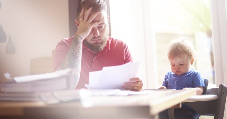 A worried-looking man sorting through papers at his dining table next to his baby in a high chair.