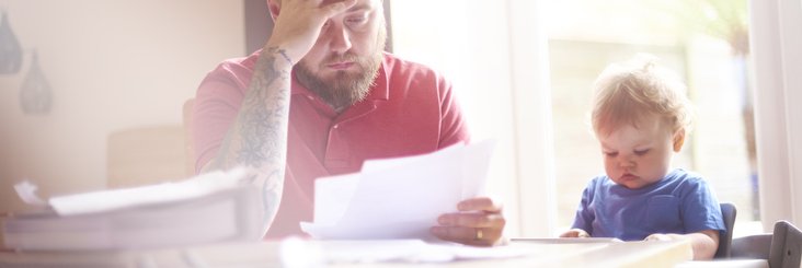 A worried-looking man sorting through papers at his dining table next to his baby in a high chair.