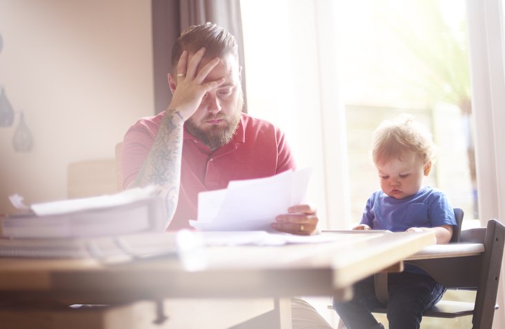 A worried-looking man sorting through papers at his dining table next to his baby in a high chair.