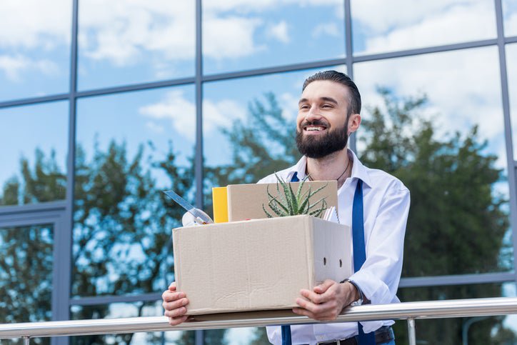 A smiling man carrying a cardboard box and walking out of an office building.