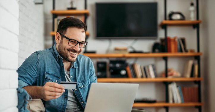 A smiling man sitting on his couch and shopping on his laptop while holding a credit card in one hand.
