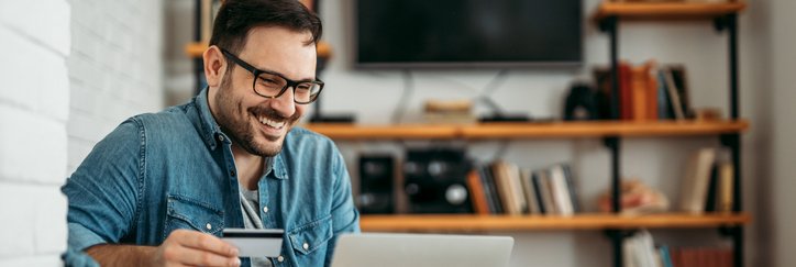 A smiling man sitting on his couch and shopping on his laptop while holding a credit card in one hand.