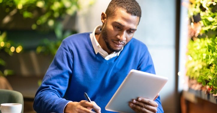 A man sitting in a cafe surrounded by green plants and writing in a notebook while holding up a tablet.
