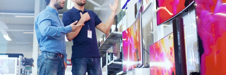 A man speaking with a salesman in an electronics store in front of a wall of TVs.