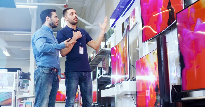 A man speaking with a salesman in an electronics store in front of a wall of TVs.