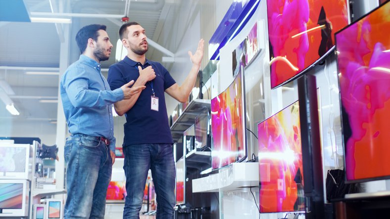A man speaks with a salesperson in an electronics store in front of a wall of televisions.
