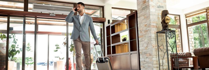 A man walking into a hotel lobby with his suitcase while on the phone.