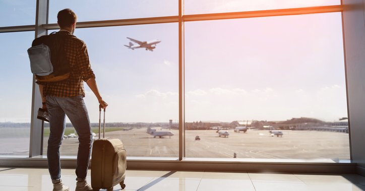 A man with a suitcase and backpack standing at an airport window and watching a plane take off