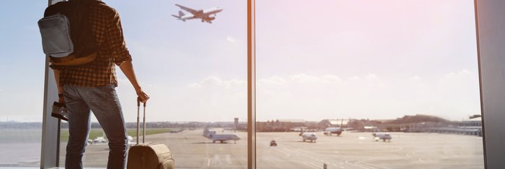 A man with a suitcase and backpack standing at an airport window and watching a plane take off