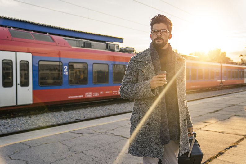A man standing on a sunny train platform with a suitcase and coffee.