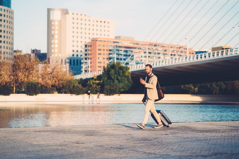 A man with a suitcase walking along a river in the middle of a city.