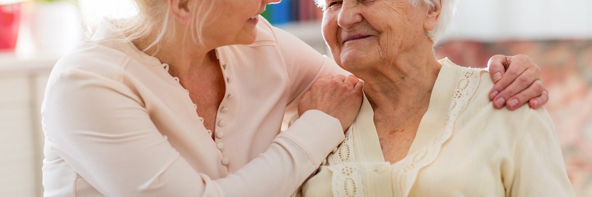 middle-aged woman holding her elderly mother and smiling while sitting at a kitchen table