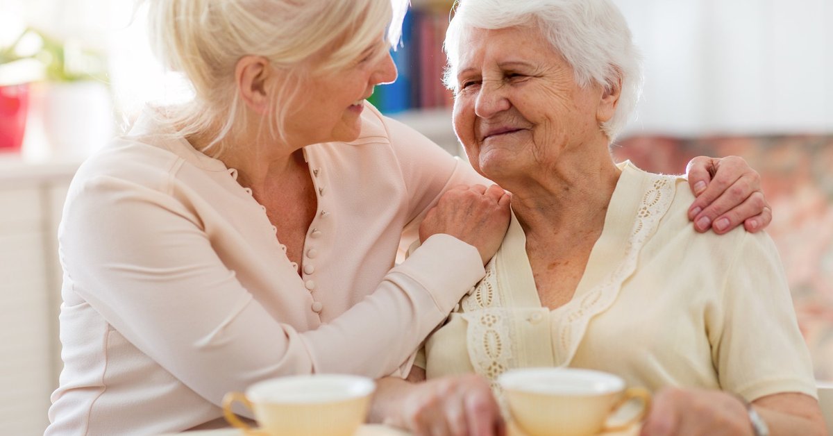 middle-aged woman holding her elderly mother and smiling while sitting at a kitchen table