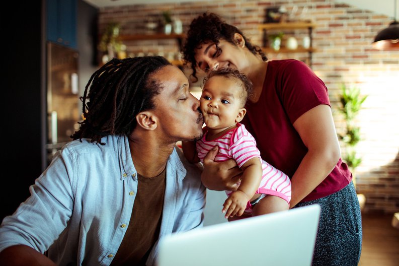 A mom holding up her baby for her husband to kiss while he works on a laptop at home.
