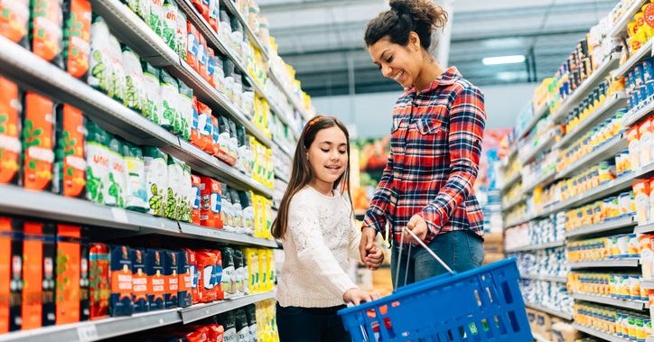 A mom and daughter walking down the aisle of a grocery and putting food in their basket.