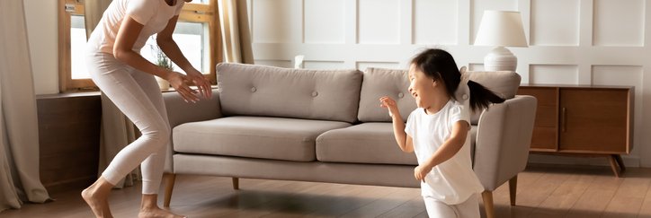 A smiling mother and her young daughter playing and chasing each other in their living room.