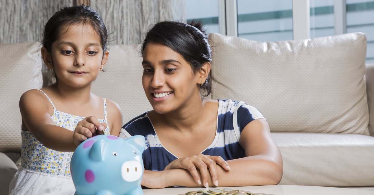 A young girl putting coins in her piggy bank as her mom looks on proudly.